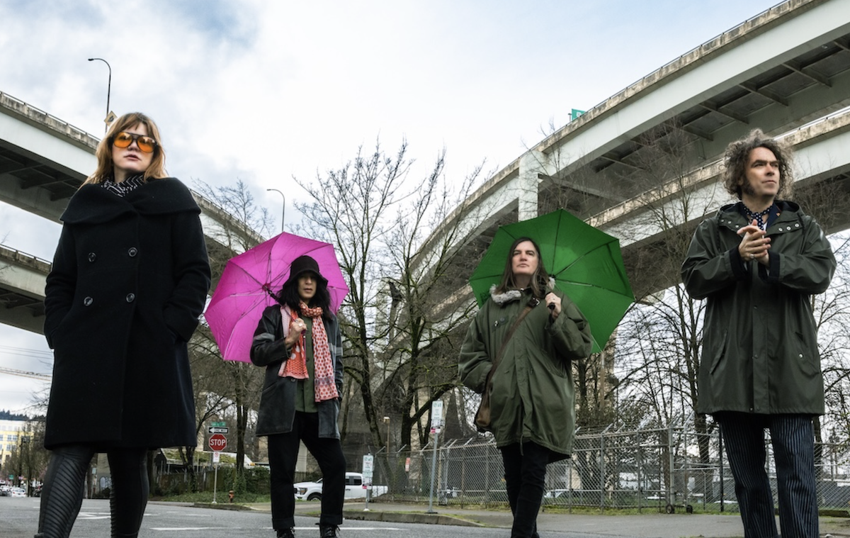 American rock band The Dandy Warhols standing outdoors near elevated overpasses with pink and green umbrellas in a press photo.